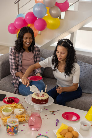 Diverse female siblings sitting on grey couch in living room, cutting red velvet cake. Celebration,...