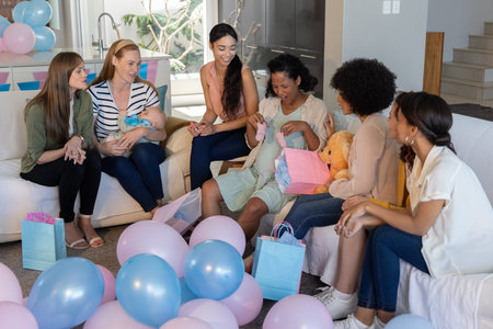 Diverse female friends opening pink baby booties in living room with pastel balloons and gift bags....