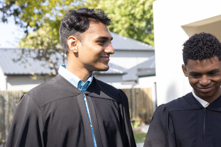 Diverse male classmates standing in yard wearing black graduation gowns over blue and white shirts....