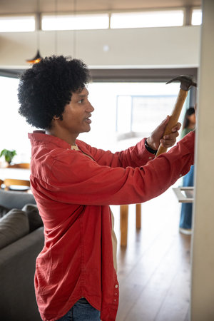 Mid adult man hammering nail into wall in living room using hammer, copy space. Home, renovation,...