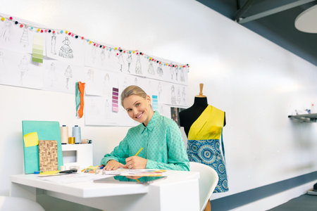 Woman sketching at design studio desk, wearing teal blouse and handling pencils and fabric. Fashion,...