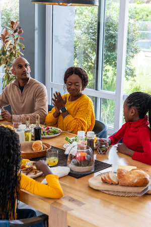Diverse family sharing meal at home dining around wooden table with mom wearing mustard sweater....