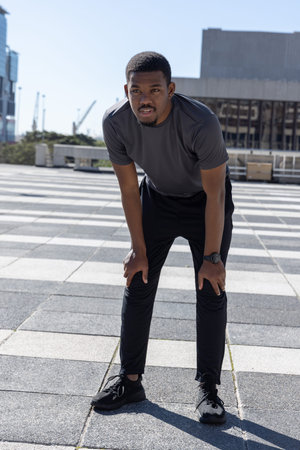 African American man leaning forward on tiled checkered plaza wearing running shoes and smartwatch. Urban, athlete, runner, sneakers, outdoor, sunlight, skylineの写真素材