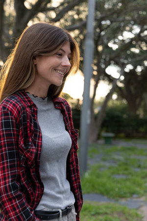 Woman standing in park during golden-hour, smiling, wearing plaid shirt with lamp post, bench....