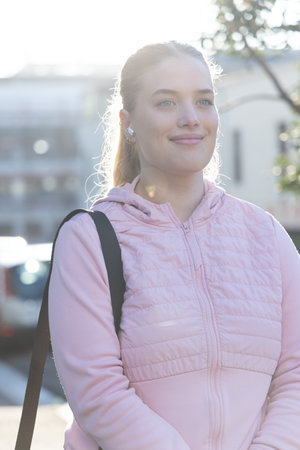 Female standing on sidewalk, wearing pink quilted hoodie, carrying black bag strap and white earbud....