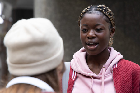 African American teenage girl talking on campus wearing red jacket backpack strap, friend in beanie....