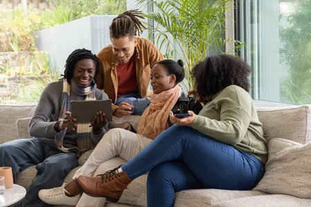 African American friends sitting on light-gray sofa at home, viewing tablet and holding remote....