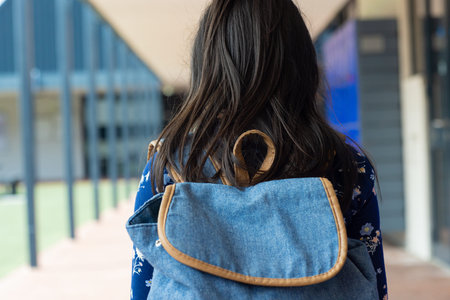 Woman with long dark hair carries a blue denim backpack, standing in a school corridor. Her floral patterned top and casual style suggest a relaxed, academic setting.の写真素材