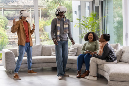 Diverse friends wearing VR headsets laughing on gray sofa in living room, one holding smartphone. Couch, lounge, group, technology, immersive, virtual, realityの写真素材