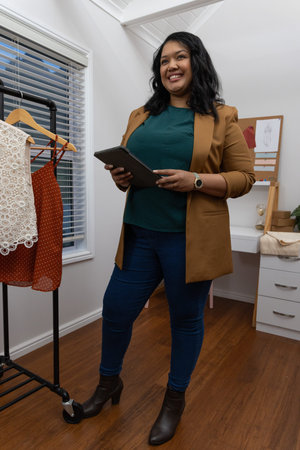 Asian woman 30s-40s in camel blazer holding tablet, viewing moodboard and rack in studio. Designer,...