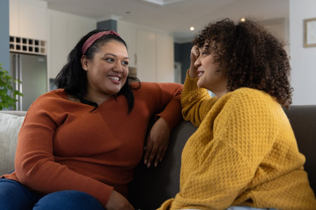 Diverse female friends chatting on sofa in open-plan living room with headband and knit sweaters....