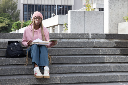 African American woman sitting on campus steps in pink hoodie, studying notebook with pen, backpack....