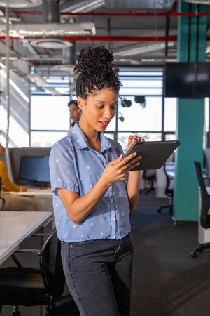 African American woman holding tablet, standing in modern office wearing blue blouse and jeans....