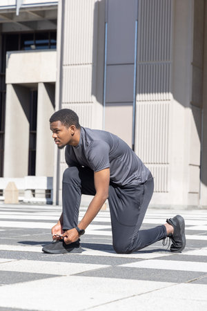 African American man kneeling and tying running shoe on patterned plaza while wearing smartwatch. Athlete, urban, outdoor, sunny, pavement, grid, architectureの写真素材