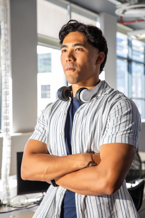 Asian man standing with arms crossed, wearing striped shirt and headphones in modern office....