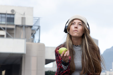 Woman holding green apple, wearing headphones, beanie and red plaid, gazing upward near building. Cityscape, urban, contemplative, fruit, earphones, cap, architectureの写真素材