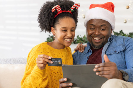 African American couple sitting on couch, wearing Santa hat and antlers, holding tablet credit-card....