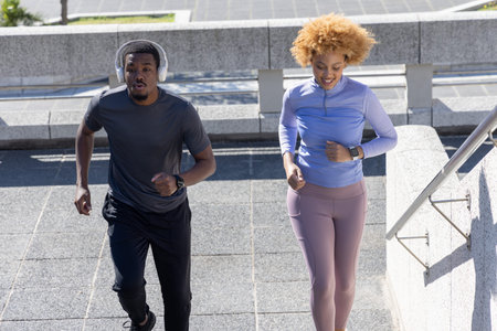 African American couple jogging up steps in urban plaza near metal handrail, wearing smartwatches....