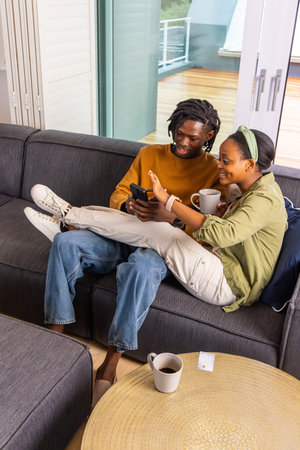 African American couple sitting closely on gray sofa at home, sharing smartphone and mugs. Couch, phone, daylight, contemporary, lifestyle, coffee, tableの写真素材