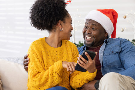 African American couple laughing and pointing at smartphone on couch at home, man wearing Santa hat. Cozy, holiday, festive, together, warmth, interior, decorの写真素材