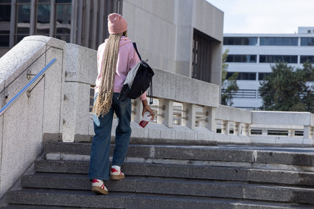 African American female climbing steps at urban campus in pink beanie and hoodie carrying backpack....