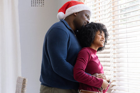 African American couple embracing at home by window with blinds, Santa hat and curtain. Romantic, partners, intimate, hug, cozy, winter, holidayの写真素材