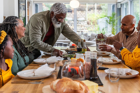 Diverse multigenerational family serving loaf of bread and pouring water pitcher at dining table. Generations, kinship, gathering, wooden, rustic, sunlit, gardenの写真素材