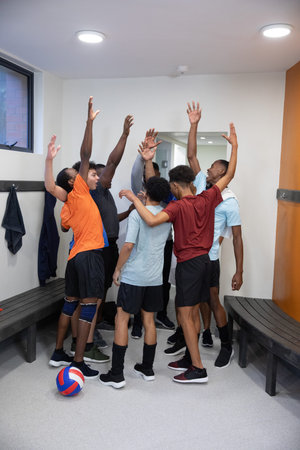 Group of eight male athletes huddling in sportswear, raising arms around volleyball in locker room....