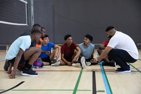 Diverse male team in sportswear sitting in arc on court, adult coach crouching, red-blue volleyball. Gymnasium, adolescent, group, training, ball, dynamic, colorfulの写真素材