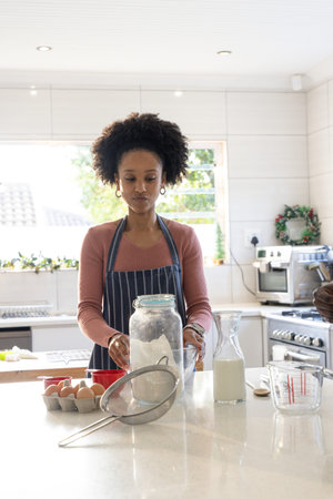 African American woman at kitchen island wearing striped apron preparing with flour jar and eggs....