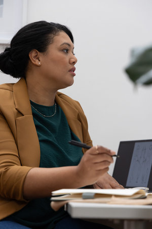 African American woman in tan blazer sitting in office at desk using laptop, notebook, pen....