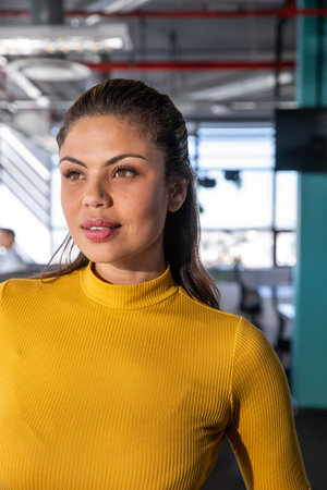 Woman in late twenties standing in modern office wearing mustard yellow turtleneck facing monitors....