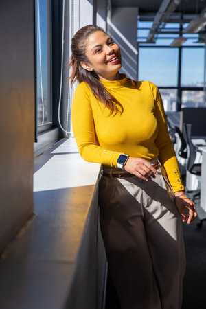 Female in 30s leaning on sunlit windowsill at office, wearing mustard top and smartwatch. Woman,...