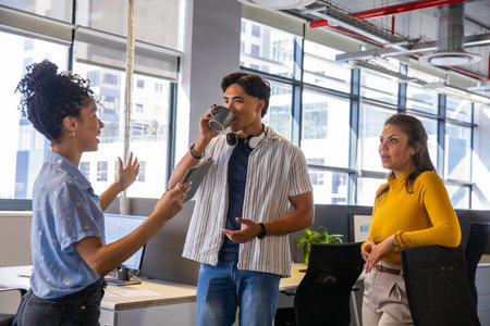 Diverse coworkers gathering near monitors in open office, gesturing, sipping mug and holding tablet. Teamwork, collaboration, discussion, meeting, professionals, colleagues, workspaceの写真素材