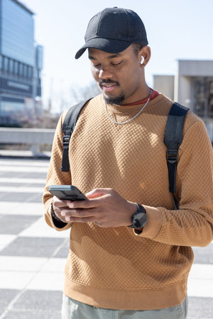 Adult African American man standing in sunlit plaza holding phone, wearing cap, backpack, earbuds....