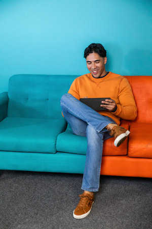 Man sitting on two-tone sofa in lounge wearing orange sweatshirt and blue jeans holding tablet. Person, device, livingroom, couch, vibrant, casualの写真素材