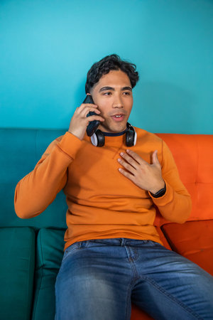 Asian man sitting on two-tone sofa in orange sweatshirt speaking on phone with headphones. Person,...