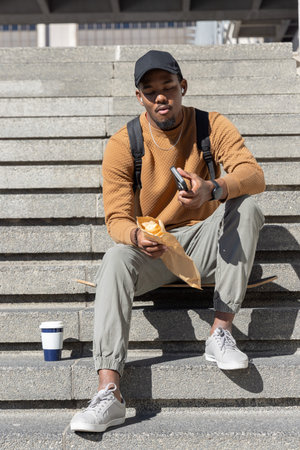 African American man 20s sitting on concrete steps in black cap and tan sweater, holding smartphone....