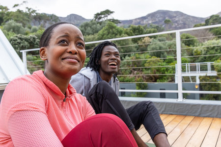 African American couple sitting on rooftop deck by cable railing on yoga mat in sportswear smiling. Outdoor, terrace, casual, athleisure, fitness, wellness, natureの写真素材
