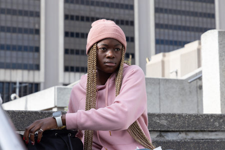 African American woman sitting on concrete steps in plaza in beanie leaning on bag with smartwatch. Urban, downtown, pink, contemplative, braids, streetstyle, cityscapeの写真素材