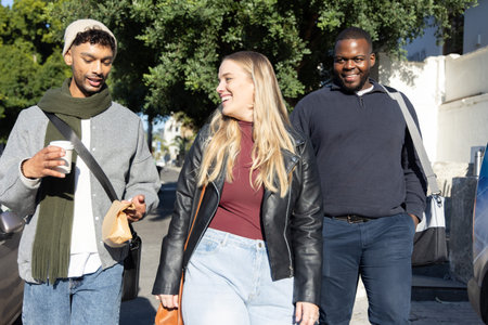 Diverse friends walking together on sunlit residential street, holding coffee cup and shoulder bags. Trio, sidewalk, urban, canopy, shade, casual, candidの写真素材