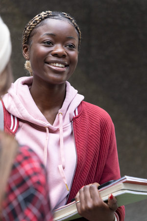 African American teenage female and peer smiling, holding books at school, wearing red jacket....