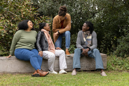 Diverse friends laughing and chatting while sitting on wooden bench in park wearing scarves. Group,...