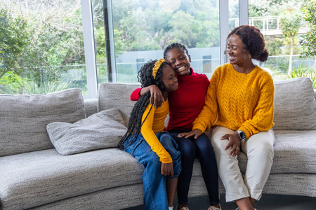African American mother with teen daughter and child sitting on gray sofa near large windows....
