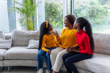 African American family sitting on sofa at home near large windows in colorful tops holding object....