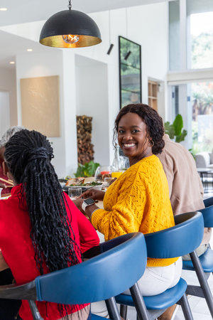 Friends sitting at countertop island in modern open-plan kitchen, woman wearing yellow knit sweater. Sunlit, dining, gathering, plants, stools, plates, blueの写真素材