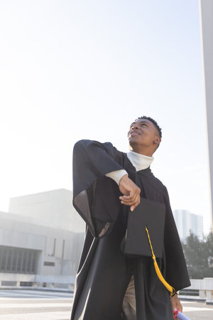 African American man standing on rooftop wearing gown holding cap with yellow tassel, copy space. Mortarboard, graduate, urban, campus, skyline, aspirational, prideの写真素材