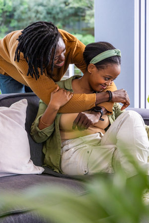 African American couple embracing in living room, man leaning over gray sofa wearing bracelet....