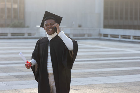 African American man standing on plaza in cap and gown holding red-ribbon diploma using smartphone. Grad, campus, outdoor, backlit, ceremony, scroll, phoneの写真素材