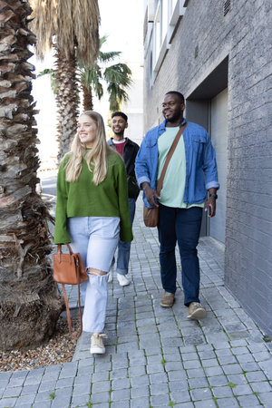 Diverse friends walking on paved path beside modern wall, woman carrying handbag, man holding phone....